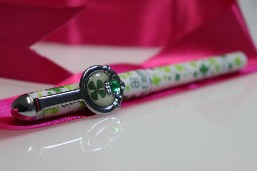 pen, lucky charm, four leaf clover, clover pen, decorative pen, green clover, pink ribbon, writing instrument, elegant gift, good luck, irish symbol, stationery, charm pen, emerald detail, shiny surface, creative gift, cute accessory, gift idea, writing, personal touch, festive decor, stylish pen, desktop item, writing tools, detailed closeup